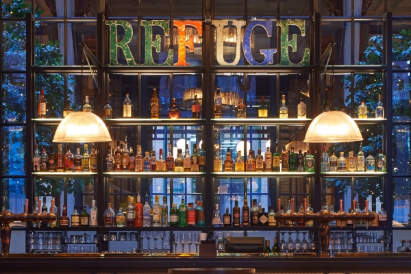 A bar with two art deco lamps hanging from the ceiling above with lots of bottles on the shelves with coloured letters spelling out REFUGE at the top